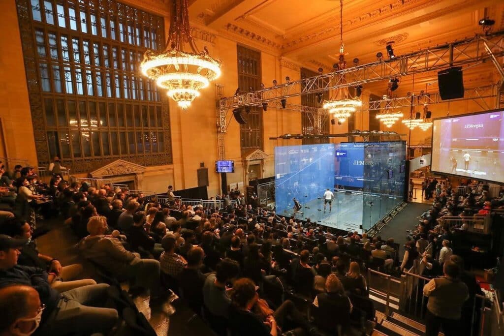 A crowd of people watching a squash match in a large hall.